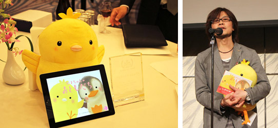 授賞式でコメントするいりやまさとし氏の写真と、展示されたぴよちゃんのぬいぐるみと電子書籍の写真です